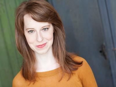 Young white woman in orange sweater with reddish hair smiling at the camera.